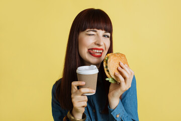 Close up of a cheerful young attractive woman grimacing at camera, showing tongue, holding tasty burger and take away coffee and enjoying her lunch time. Isolated on yellow studio backgroud