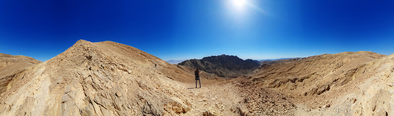 Fototapeta na wymiar male hiker on a hiking trail in Eilat mountains. Colorful nature pattern painted on a rock wall. Rock formations and boulders. Panoramic view over the trail on surrounding red mountains