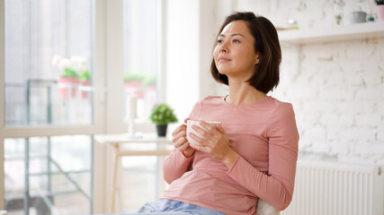 Multi ethnic satisfied asian female enjoying cup of coffee or tea drinking warm beverage at home. Young girl relaxing with morning drink in hands looking through window enjoying and dreaming