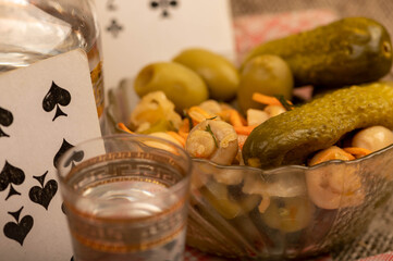 Playing cards, a glass of vodka, a decanter of vodka and a glass dish with pickles and mushrooms on the table.