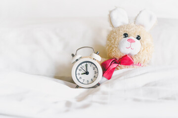 Cute little rabbit doll lying alone with analog alarm clock  on white bed for wake up or go to bed,...