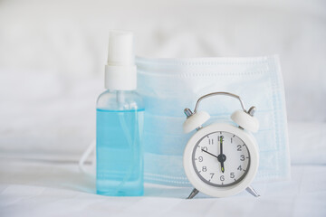 white analog clock , blurred alcohol spray bottle and medical mask on bed , new normal lifestyle after loosen lockdown because coronavirus (covid-19)
