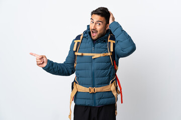 Young mountaineer man with a big backpack isolated on white background surprised and pointing finger to the side