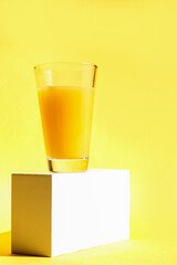 A glass of freshly squeezed refreshing orange juice on a white pedestal or podium. Yellow background. The concept of a healthy lifestyle, raw food diet, vegetarianism. Vertical orientation. Copy space