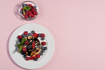 top view of homemade American pancakes with fresh blueberries, raspberries, strawberries and chocolate syrup. Healthy breakfast in the morning. Pink background
