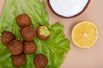 Close up with fried falafel on salad leaf, white yogurt sauce in clay bowl and fresh yellow lemon on beige background. Healthy roasted vegetarian fastfood. Israeli cuisine.