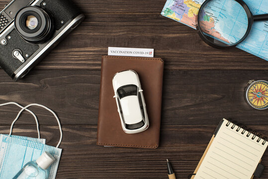 Top view photo of car model on leather passport cover with vaccination certificate camera map magnifier compass notebook pen medical masks and sanitizer on isolated wooden table background