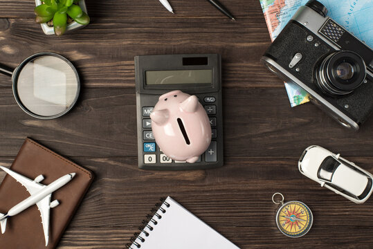 Top view photo of piggy bank on calculator in the center plane and car models camera map magnifier compass notebook passport cover and plant on isolated wooden table background