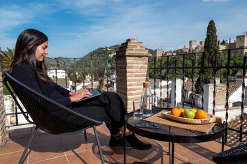 Female travel blogger working on laptop while sitting on terrace with view of Alhambra fortress in Granada, Spain