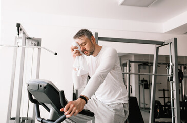a tired man wipes sweat from his face with a towel during an exercise on an exercise bike.