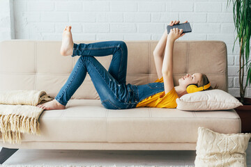 Teenage girl in denim overalls relaxes in pleasure on the couch with an electronic tablet. The...