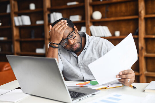 Displeased African-American man looking through workpapers, stressed black male employee reading a document bank statement sitting in the office, has bad balance, bankruptcy, debt