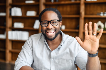 Headshot of happy African-American guy wearing eyeglasses looking at the camera and waving hello, video chat with male colleague or teacher, a mixed-race man holding video meeting, online conference