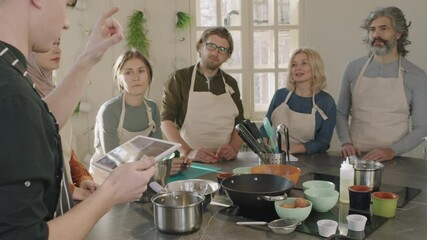 Medium shot of group of diverse people listening to young male chef with digital tablet, learning cooking techniques at culinary master class