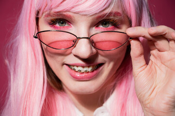 Close-up portrait of a young woman with braces in a pink wig and sunglasses on a pink background.