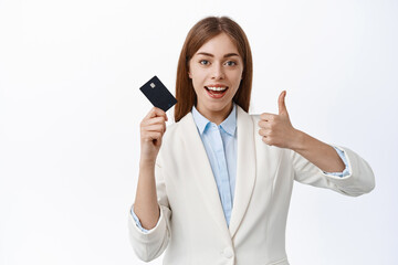 Fototapeta premium Cheerful girl office manager in business suit, shows plastic credit card and thumbs up, approve bank service, recommends product, standing over white background