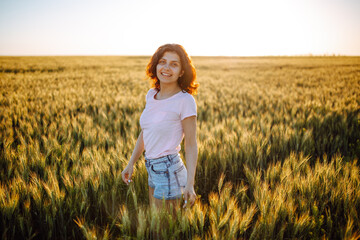 Smiling girl stands in the field. Happy woman at sunset in wheat. A girl in a pink T-shirt stands against the background of the field and the setting sun.