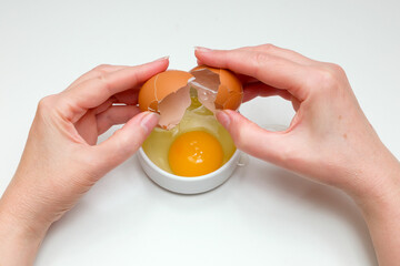 women's hands breaking a raw chicken egg into a small white cup on a white background
