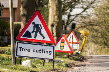 Red and white tree cutting warning signs standing at side of road with road works worker symbol.  Traffic sign alerting drivers of danger.  Traffic lights and road narrow distance, traffic management