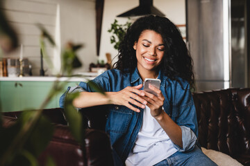 Relaxed young african woman texting on her phone in the modern house.Smiling african american woman using smartphone at home, messaging or browsing social networks while relaxing on couch - Powered by Adobe