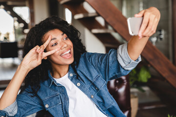 Happy afro young woman taking selfie on her phone in the living room. African businesswoman in home office. Beautiful young women making selfie photo in the home office.