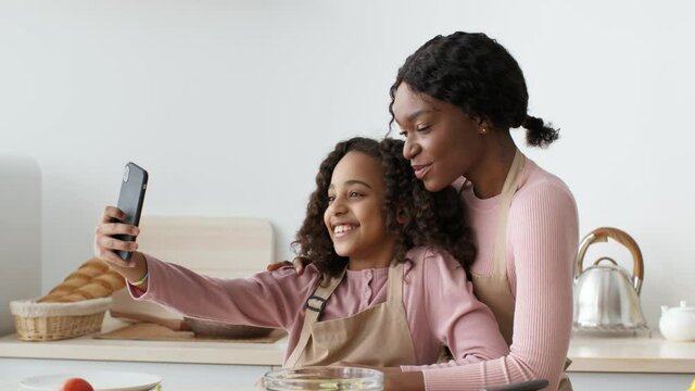 Social media content. Happy african american girl taking selfie with her mother, smiling to smartphone at kitchen