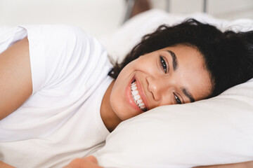 Fototapeta na wymiar Happy smiling young african woman lying in her white bed in the morning. Young African American woman waking up at home.