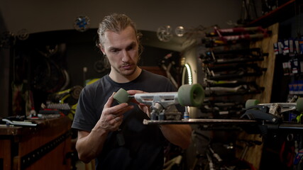 Young Man In The Workshop Attaches Wheel To Skateboard. Many Tools And Parts In The Workshop. Handsome Guy With Beard Works Carefully.