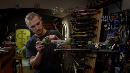 Young Man In The Workshop Attaches Wheel To Skateboard. Many Tools And Parts In The Workshop. Handsome Guy With Beard Works Carefully.