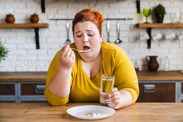 Fat plus size obese young woman sadly eating pills for losing weight in the kitchen. Healthy...