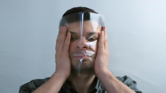 Portrait of young man is puttion on plastic transparent film on his face on white background. He is looking at camera. He makes terrible faces and fools around.
