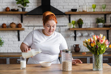 Young chubby woman standing in kitchen pouring milk into oat-flakes smiling cheerful while cooking breakfast. Fat woman on a diet. Proper nutrition.