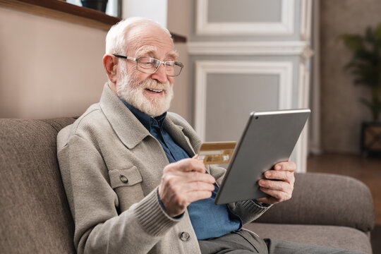 Cheerful old grandfather man doing online payment from digital tablet with credit card. Elderly senior caucasian grandpa paying bills online with credit card