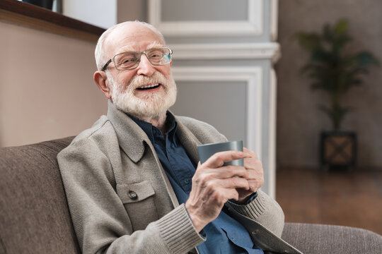 Delicious Drink. Pleasant Elderly Man In Spectacles Sitting On The Sofa And Posing With A Cup Of Coffee While Smiling At The Camera. Laughing Elderly Man Sit Relax On Couch In Living Room Drinking Tea