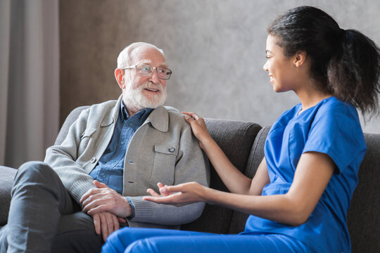Caring african-american nurse talks to old elderly senior patient holds his hand sit in living room at homecare visit provide psychological support listen complains showing empathy encouraging.