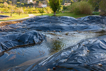 Black plastic awnings covering the side of a river to prevent the growth of invasive reeds (Arundo Donax).