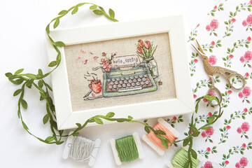 close up cross stitch typewriter saying hello spring on linen canvas in white frame, floss threads...