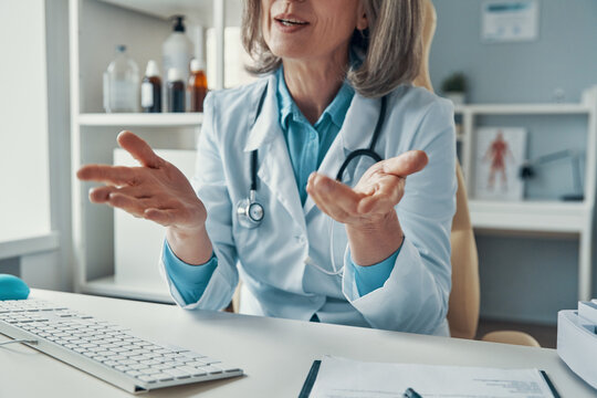 Close up of mature female doctor in white lab coat telling something and gesturing while sitting in her office
