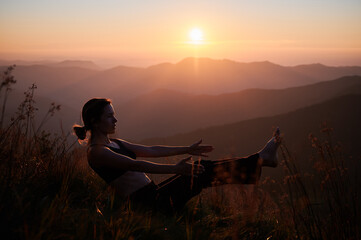 Beautiful young woman in sportswear performing yoga pose on grassy hill with yellow orange sky on background. Fit woman practicing yoga and working out in the mountains.