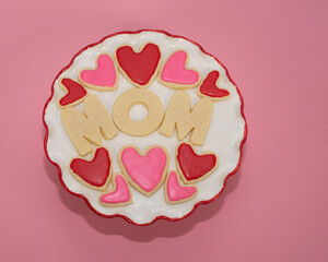 Mother's Day is celebrated with homemade sugar cookies spelling MOM and pink and red heart shaped cookies on a round white and red plate against a pink background.