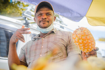 young dark-skinned man of Haitian nationality working selling fruit on the street with a bicycle and an umbrella and wearing a mask. selling pineapple - obrazy, fototapety, plakaty