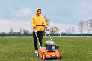 Positive handsome guy in yellow hoodie with gasoline mower in his hands, man mows grass on lawn, improvement of territory, male trimming field with grass-cutter.