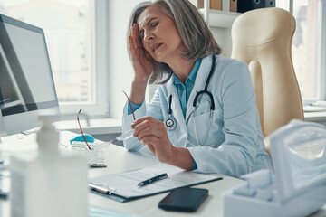 Fototapeta na wymiar Overworked mature female doctor in white lab coat suffering from headache while sitting in her office