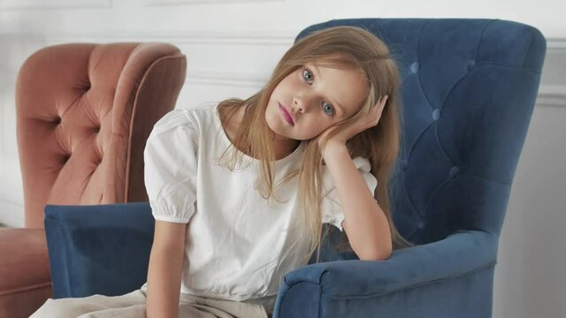 Close up portrait of cute serious  little blonde caucasian girl model with big eyes looking at camera. Portrait of adorable preschool american kid child sitting on armchair. Concept of children