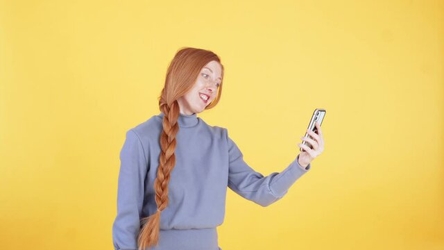 Young woman with freckles and long ginger plait is talking to someone on the phone using video call and smiling. Yellow background
