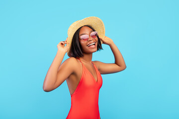 Portrait of lovely black lady in straw hat and swimsuit posing and smiling at camera on blue background