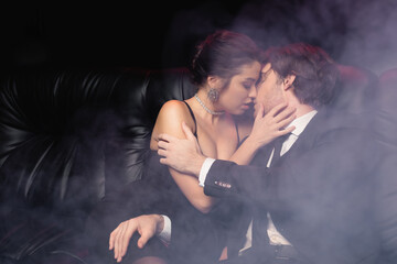 passionate woman in slip dress kissing man in suit on black with smoke.
