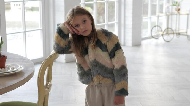 Close up portrait of serious little caucasian girl looking at camera. Portrait of adorable preschool kid in fur coat standing and leaning on chair in front of windows at home
