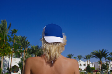 Young beautiful girl with blonde hair in a blue and white trucker hat on a background of blue sky and palm trees, baseball cap mockup.