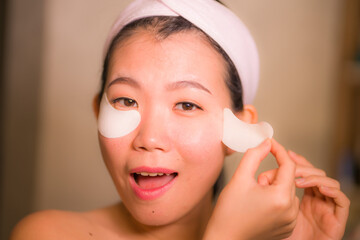 portrait of young happy and beautiful Asian Chinese woman applying facial anti panda eyes patch at bathroom mirror in skin care and beauty treatment concept